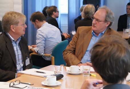 Teilnehmer sitzen am Gruppenarbeitstisch und sprechen miteinander. Im Hintergrund sitzen weitere Teilnehmer an Gruppenarbeitstischen.