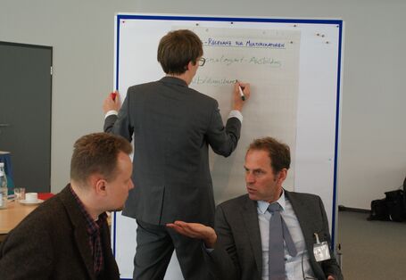 Teilnehmer schreibt auf Papier an Moderationswand. Im Vordergrund sitzen zwei Teilnehmer im Gespräch.