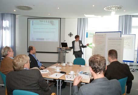 Teilnehmer sitzen am Arbeitgruppentisch und hören der Vortragenden zu. Die Vortragende spricht zu den Teilnehmenden und zeigt auf Themen an Moderationswänden.