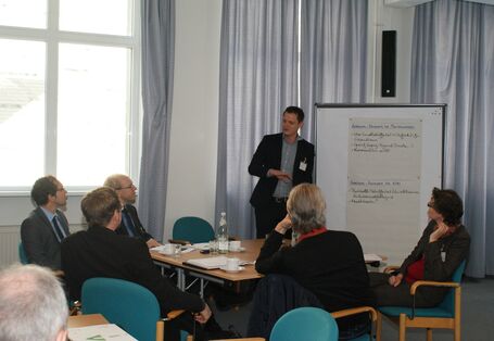 Teilnehmer sitzen am Gruppenarbeitstisch und hören Vortragenden zu. Der Vortragender steht neben einer Moderationswand.