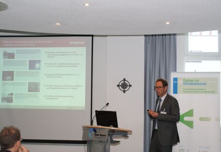 Vortragender steht am Stehpult. Im Hintergrund ist der Vortrag auf Projektionswand und Veranstaltungsaufsteller zu sehen.