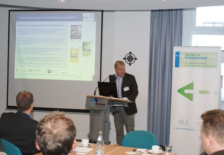 Vortragender steht am Stehpult. Im Vordergrund sitzen Zuhörer und im Hintergrund ist der Vortrag auf der Projektionswand und der Veranstaltungsaufsteller zu sehen.