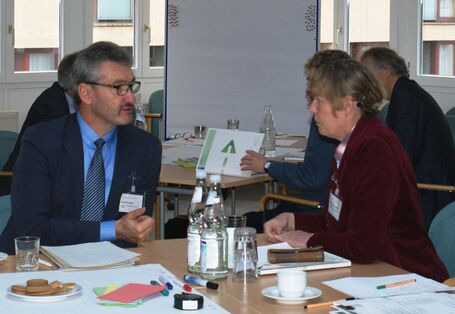 Zwei Teilnehmer befinden sich im Dialog miteinander sitzend am Gruppenarbeitstisch. Im Hintergrund stehen weitere Gruppenarbeitstische vor einer hellen Fensterfront.