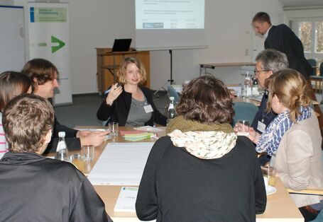 Teilnehmer sitzen am Gruppenarbeitstisch und hören einer Teilnehmerin bei ihrem Redebeitrag zu. Im Hintergrund sieht man den Veranstaltungsaufsteller, das Rednerpult und die Projektionswand und einen anderen Teilnehmer stehen.