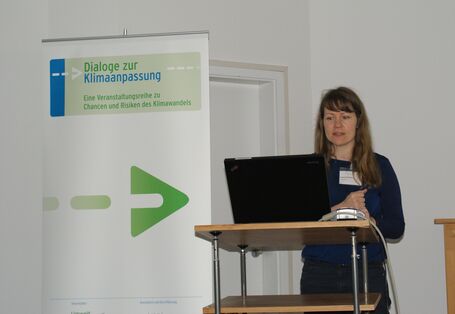 Teilnehmende steht am Rednerpult vor geöffnetem Laptop. Im Hintergrund steht der Veranstaltungsaufsteller.