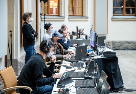 Die Technikerinnen und Techniker sitzen in einer langen Reihe und schauen konzentriert in die Laptops. Sie tragen Kopfhörer und Mund-/Nasenbedeckungen.