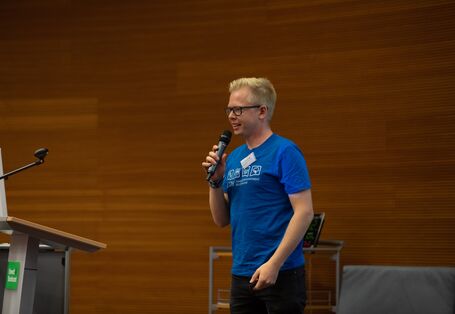 Ein Vortragender spricht in ein Mikrofon, welches er in der Hand hält. Der Vortragende trägt ein blaufarbenes T-Shirt und eine Brille.