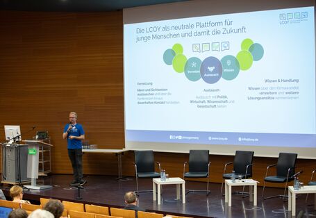 Ein Vortragender steht vor einer großen Leinwand auf dieser seine Präsentation abgebildet ist. Die Präsentation stellt mittig ineinander verschränkte Kreise in den Farben Blau und Grün dar.