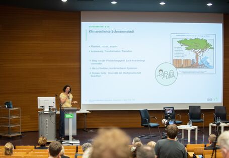 Der Blick geht vom Publikum in Richtung Bühne auf die große Leinwand die eine Präsentation mit der Überschrift: Klimaresiliente Schwammstadt zeigt. Davor steht eine Vortragende und spricht in das Mikrofon. Sie trägt ein gelb weiß geringeltes Shirt.