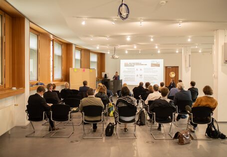 Der Blick geht von hinten über die sitzenden Workshopteilnehmden hinweg nach vorne auf die Leinwand mit einer Präsentation. Die Vortragende steht am Stehpult und stellt Ihre Präsentation vor. Linke Seite entlang zeigt das Bild eine lange Fensterfront.