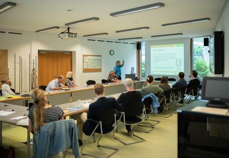 Workshopteilnehmende sitzen in einem hellen Raum an einer rechtigen Tischformation. Der Blick geht hinaus zum Fenster. Vor dem Fenster steht die Präsentationsleinwand. Der Vortragende zeigt auf seine Präsentation. Er trägt ein hellblaues Shirt.