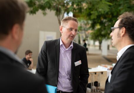Drei Teilnehmende in dunkler Kleidung unterhalten sich miteinander. Im Hintergrund sind ein Baum und Konferenzmaterialien zu sehen.