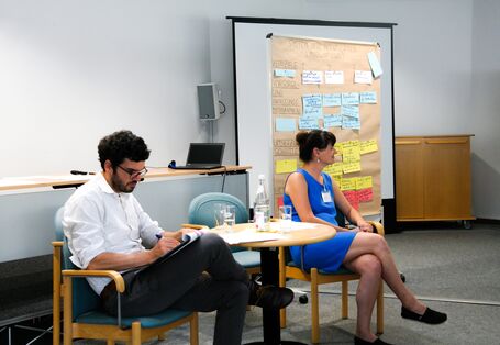 Reflexion: Die Durchführenden von der Veranstaltung sitzen zu zweit an einem Tisch. Sie schauen und hören in die Runde. Im Hintergrund steht eine Moderationswand, die mit diversfarbigen und beschrifteten Moderationskarten bestückt ist. Die Frau trägt ein blaues Sommerkleid. Der Mann ein helles Hemd und schwarze Hose.