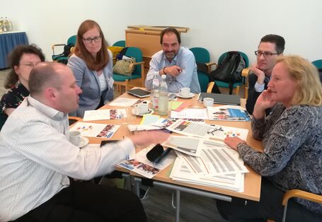 Teilnehmende sitzen am Gruppenarbeitstisch und hören einen Redner interessiert zu. Auf dem Tisch liegen Arbeitsunterlagen und es stehen Getränkeflaschen und Gläser drauf. Auf den umliegenden Stühlen sind Taschen und Jacken abgelegt.