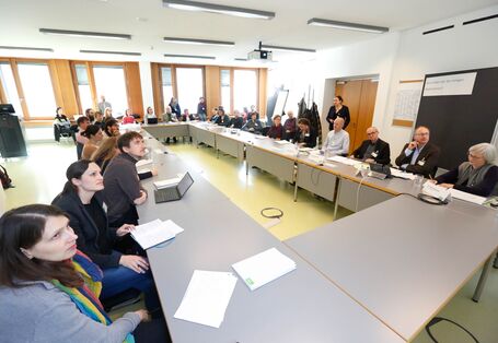 Teilnehmende sitzen an hellgrauen Tischen im Rechteck und schauen hoch zur Präsentationsfläche.