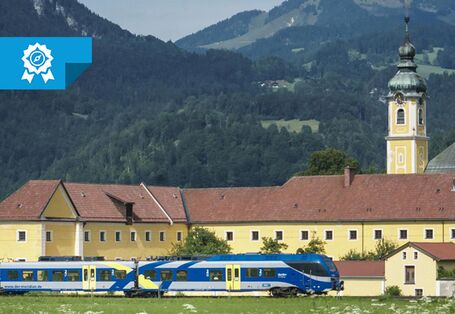 Im Vordergrund fährt ein Personenzug vor einem größen, gelb gestrichenen Gebäude vorbei. Zu dem Gebäude gehört ein Turm, der rechts im Bild zu sehen ist. Im Hintergrund sind Berge erkennbar. Links oben über dem Bild liegt ein Banner des Blauen Kompasses.