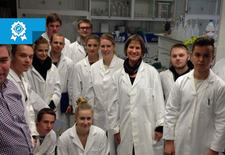 Gruppenbild aus dem Labor. Personen tragen weiße Kittel. Im Hintergrund befinden sich Labormaterilien in und außerhalb von verglasten Laborschränke. Eine Mann im linken Vordergrund trägt Alltagskleidung: Hose und Hemd.