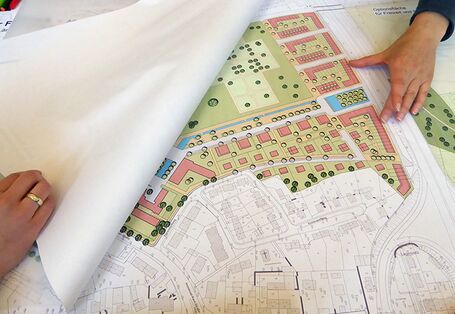 Die Rahmenplanung liegt aufgeschlagen vor. Zwei Hände halten das Papier aufgeschlagen und zeigend auf die farbige Projektskizze.