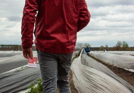 Fotografie einer Person auf einem bewirtschafteten Feld. Nur der Rumpf ist erkennbar, in der Hand hält die Person einen Flyer.