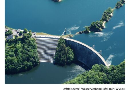 Blick von oben auf eine Talsperre. Treppen führen hinab auf der Seite mit niedrigerer Wasserhöhe. Grün belaubte Bäume auf einem Wall im Wasser und drum herum sowie Gebäudeteile auf der Talsperre runden das Bild ab. Das Wasser erscheint in einem tiefen klaren Blau.