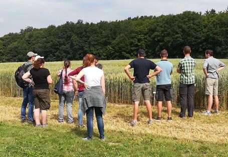 Auf einem Acker stehen mehrere jüngere Personen mit dem Rücken zur Kamera. Eine verdeckte Person hält einen Vortrag, dem die anderen zuhören.