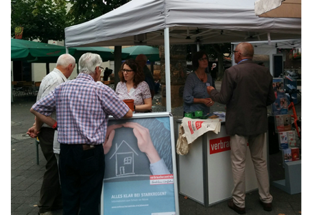 Mehrere Personen stehen um und hinter einem Marktstand der Verbraucherzentrale NRW mit heller Überdachung. Im Vordergrund ein Aufstellplakat, auf dem sich schützend eine Hand über ein mit Kreide gezeichnetes Haus hält.