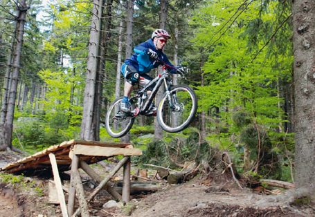 Ein hölzerner Weg mit Absprungfläche für Mountain Biker verläuft im Wald als Downhill-Strecke im Fichtelgebirge. Im Vordergrund passiert ein Mountainbiker gerade die Absprungläche.