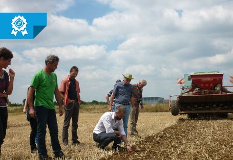 Mehrere Personen stehen auf einem Feld. Im Hintergrund fährt ein Mähdrescher. Einige der Personen begutschten den Boden.