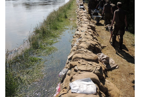 Fotografie eines Sandsackwalls. Links des Walls ist Wasser, aus dem überschwemmte Gräser herausragen, rechts gehen und stehen einige Personen mit Schubkarren auf einem Weg.