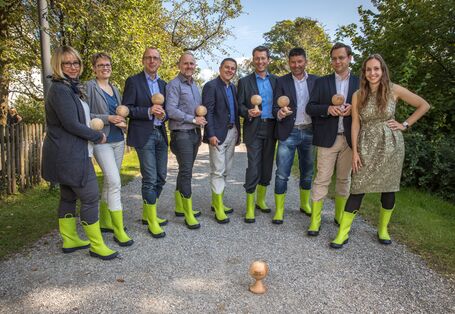 Teilnehmer/-innen der Pilotschulung stehen im Halbkreis auf einem Kiesweg vor blauem Himmel und belaubten Bäumen. Sie halten ein hölzerne pokalähnliche Form in Händen, eine Form steht auf dem Boden vor dem Halbkreis. Die Teilnehmer/-innen tragen hellgründe Gummistiefel.