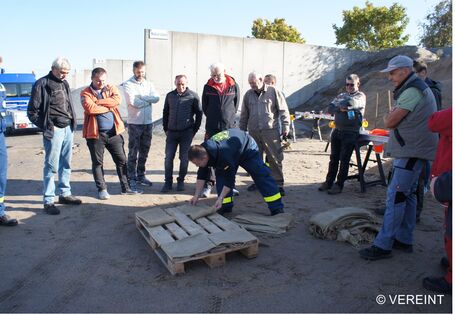 Eine Menschentraube schaut einem THW-Mitarbeitenden dabei zu, wie er das Sandsackstapeln auf einer Europalette demonstriert.