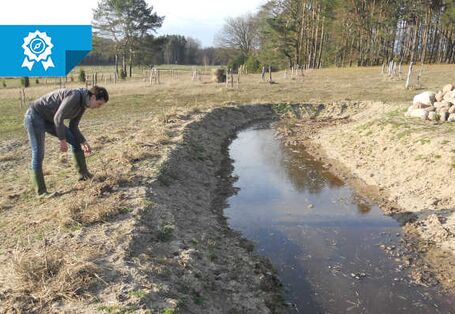 Eine Person beugt sich über einen Wassergraben, der durch ein Feld geht.