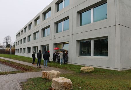 Vor einem grauen Gebäude stehen mehrere Personen in einer Gruppe um ein Messgerät.