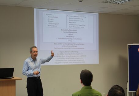 Vortragender steht gestikulierend vor Publikum. An seiner linken Seite stehen zwei Moderationswände, an seiner rechte Seite befindet sich ein Rednerpult. Auf dem Rednerpult liegt ein aufgeklapptes Laptop.