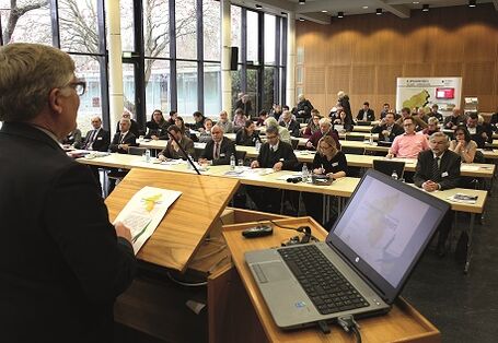 Vortragender steht am Pult bei geöffnetem Laptop vor den Teilnehmern, die in Reihen an Tischen sitzend zuhören.