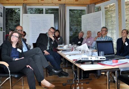 Workshopteilnehmer und -teilnehmerinnen sitzen um einen Gruppenarbeitstisch umgeben von Panoramafenster und schauen auf die Flipcharts mit den handschriftlichen Präsentation der erarbeiteten Ergebnisse