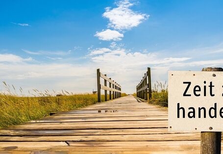 Auf einem Schild im Vordergrund steht Zeit zu handeln. Im Hintergrund führt ein Holzdünenweg optisch in den Himmel hinein.