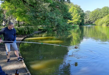 Gewässeruntersuchung am Waller Park See Bremen: ein Mann steht mit einer langen Stange am Wasser vor grünbelaubten Bäumen.