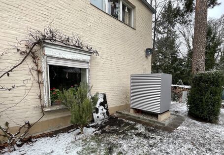 Ein Außengerät einer Wärmepumpe im leicht verschneiten Garten, quer zur beigen, geziegelten Hauswand aufgestellt.