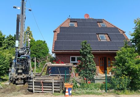 Ein geklinkertes Haus steht quer zur Straße, das Dach ist mit Photovoltaik belegt. Vor dem Haus im Vorgarten steht ein hohes Bohrgerät, daneben Stangen auf einer Palette und ein Container.