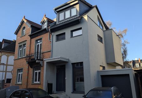 Die Fassade eines zweigeschossigen, rot geklinkerten Altbaus mit einem modernen, grau verputzten Anbau.