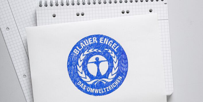 Foto von oben. Auf einem Schreib-Block liegt ein weißes Blatt Papier. Darauf ist das runde, blaue Logo vom „Blauen Engel“ mit blauem Stift gemalt. Ganz oben liegt ein zusammengeknülltes Stück Papier.