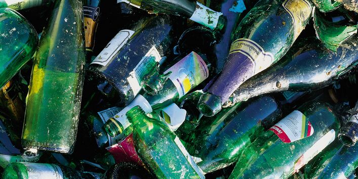 A high-angle, close-up shot of a pile of discarded green and dark glass bottles intended for recycling. The bottles, mostly wine and champagne varieties, are cluttered together, some broken, with visible paper labels and residue, showing textures of dirt and wear.