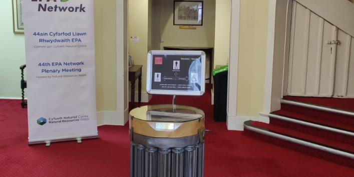 Entrance hall with a red carpet. In the foreground a monitor displaying info and on the left a rectangular poster announcing a meeting.
