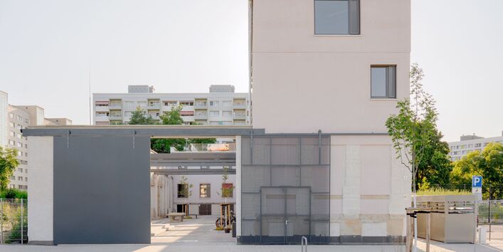 Frontalansicht eines Hofeingangs. Rechts ein dreigeschossiges Gebäude mit heller Fassade, Flachdach und unregelmäßig angeordneten Fenstern. Auf der linken Seite eine geschosshohe Einfassung des betonierten Innenhofs, in den durch das geöffnete graue Schiebetor der Blick fällt. Im Hintergrund ein Plattenbau.