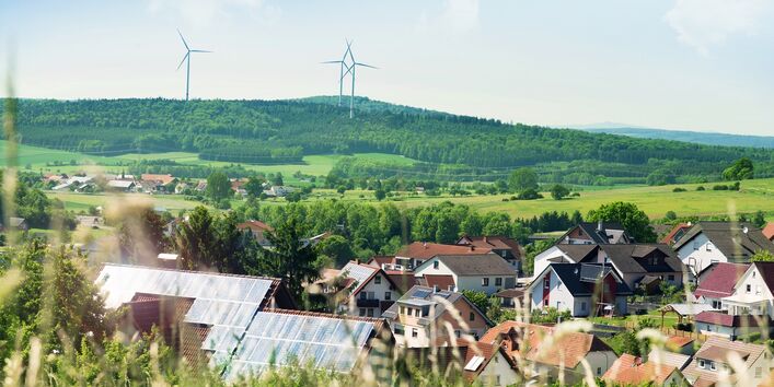 Häuser mit Photovoltaik auf dem Dach, in der umliegenden hügeligen Landschaft Windenergieanlagen