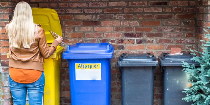 Eine Person mit blonden Haaren steht von hinten gesehen vor vier verschiedenen Mülltonnen (gelb, blau, zwei dunkelgraue) an einer Ziegelwand. Die Person öffnet gerade den Deckel der gelben Tonne, um eine leere Medikamenten-Blisterpackung einzuwerfen. Auf der blauen Tonne ist die Aufschrift „Altpapier“ zu erkennen.