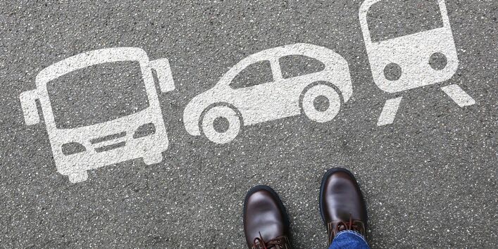 Auf den Asphalt sind Piktogramme eines Busses, eines Autos und eines Zuges gesprüht. Davor steht ein Mensch, von dem nur die Schule zu sehen sind. 