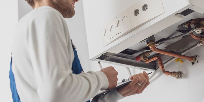 Foto. Ein Handwerker in blauer Arbeits-Hose steht vor einer weißen Gas-Therme an der Wand. Er schaut auf die Rohre unter dem Gerät und schreibt etwas auf ein Klemm-Brett.
