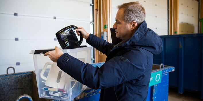 Foto. Ein Mann in einer blauen Winter-Jacke steht vor einem großen, blauen Müll-Container. Er hält eine durchsichtige Kiste aus Plastik. Darin liegt ein weißer Wasser-Kocher. Er legt ein schwarzes Elektro-Gerät in den Container.